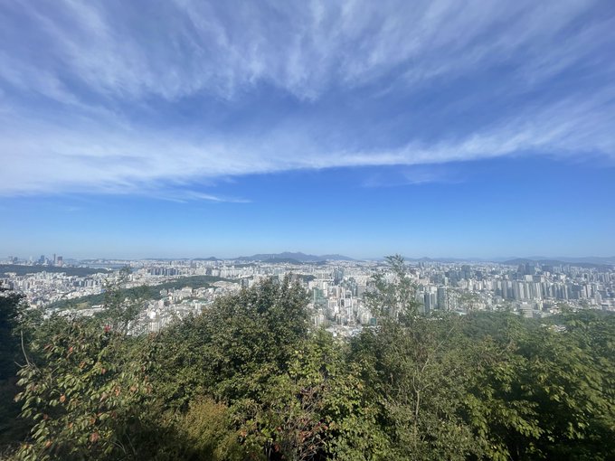 A scenic panoramic photograph showing a vast cityscape under a bright blue sky with wispy clouds, viewed from a lush green hillside, with distant mountains on the horizon.