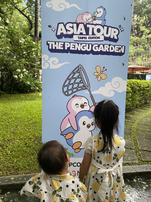 Two young children in matching yellow-and-black polka-dot dresses stand before a blue promotional banner reading 'ASIA TOUR TAIPEI EDITION - THE PENGU GARDEN' featuring cartoon penguin characters with a butterfly net.