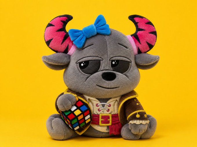 A photograph of a gray plush toy with pink and black striped horns and a blue bow, holding a Rubik's cube and wearing a small outfit with a belt and dagger, set against a yellow background.