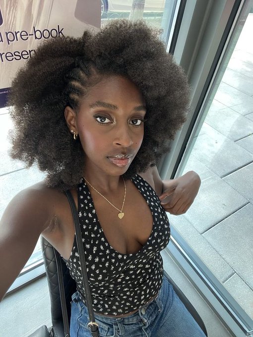 A young woman with voluminous natural curls takes a selfie wearing a black floral crop top and delicate gold heart necklace, her serene gaze capturing an ethereal beauty against a windowed backdrop with partial text reading "pre-book" and "resen." The softly lit portrait and graceful pose perfectly embody the "ethereal" essence of the post.