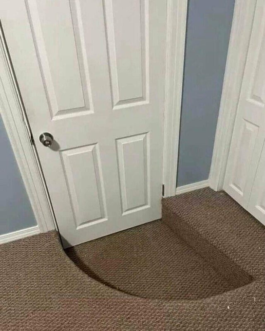 A white interior door shown with a large section of carpet peeled up from the floor, creating a visible gap and fold on the brown carpeted surface.