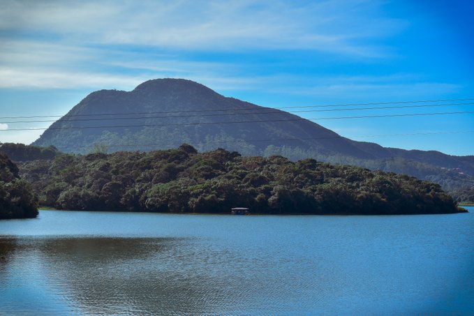 A scenic landscape photograph showing a calm lake with a forested island and a large mountain under a blue sky with light clouds.