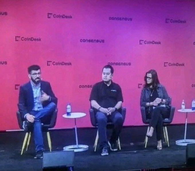 Three panelists sit on stage at a CoinDesk Consensus conference, seated in front of a red backdrop featuring repeating "CoinDesk" and "Consensus" logos, with water bottles placed on small tables between them as they engage in a discussion.