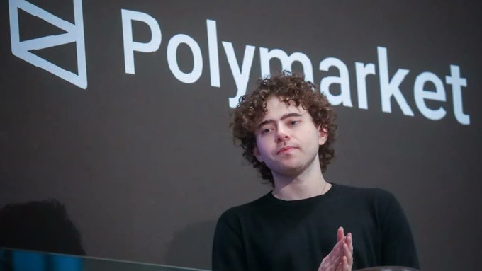 The image shows a man with curly hair standing next to a large screen displaying the Polymarket logo, which reads "Polymarket" in white letters against a dark background. The man appears to be giving a speech or presentation at an event, possibly related to cryptocurrency trading or blockchain technology.