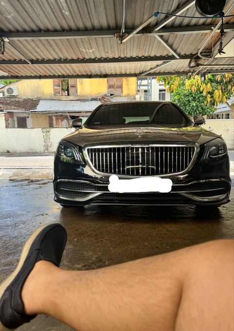 A black luxury sedan parked under a covered area with someone's leg and black slip-on shoe visible in the foreground.