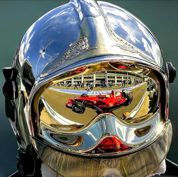 A highly reflective chrome/silver racing helmet whose mirrored visor reflects a red Formula 1 race car on a track.