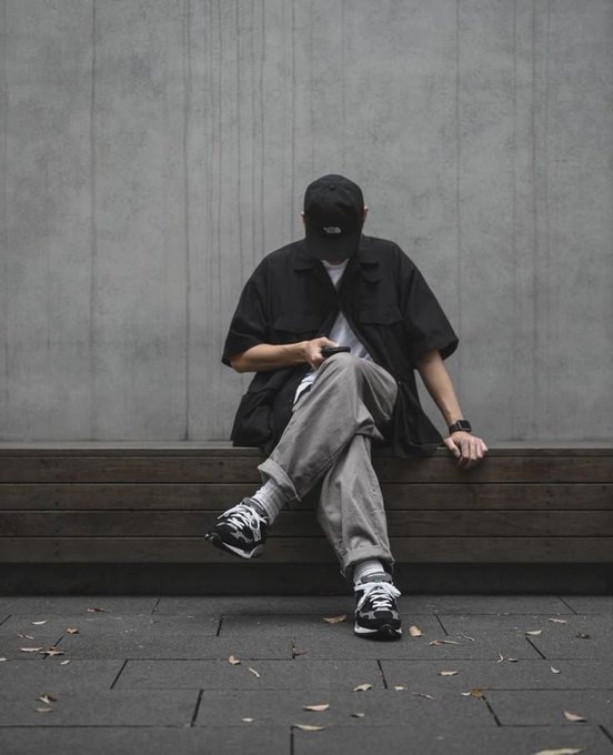 A person in a black cap, black short-sleeved shirt, and gray pants sits on a wooden bench against a concrete wall, looking down with their cap obscuring their face.