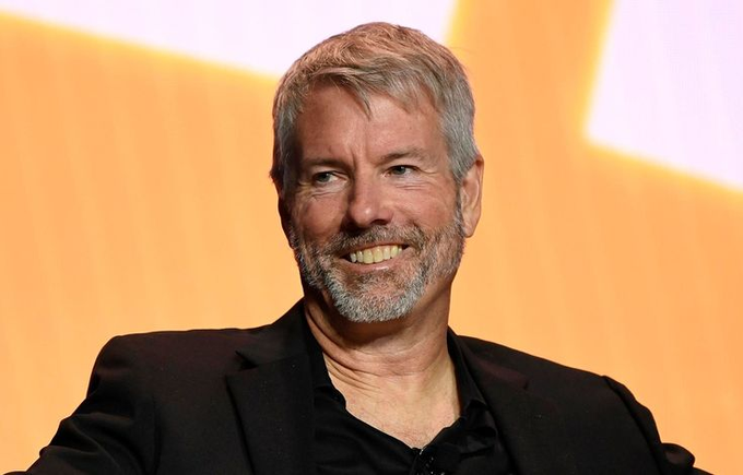 A portrait of a smiling older man with gray hair and a beard, wearing a black suit jacket over a black shirt, posed against an orange background.