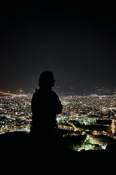 A silhouetted figure wearing a hoodie stands on a hill overlooking a sprawling, brightly lit city skyline at night.