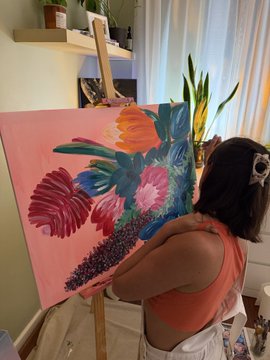 A woman with dark hair wearing a coral top is painting a colorful tropical floral artwork on a pink canvas in an art studio.