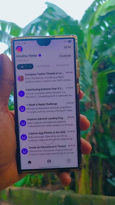 A hand holds a smartphone displaying the Gibwork platform profile "Umainz Forex" (ref: akong_jnr) showing a balance of $2.54 with 15 referrals, alongside various available tasks and bounties ranging from $100 to $400 such as "Compose Twitter Thread or Lo…" and "Yield During Extreme Fear X" — illustrating the user's progress on Day 21 of their quest to get Bybit to create a contest on Gibwork.