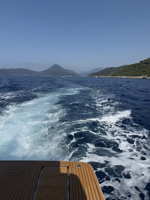 A scenic view from the back of a yacht showing the boat's wake on blue ocean waters with green mountainous coastline under a clear sky.
