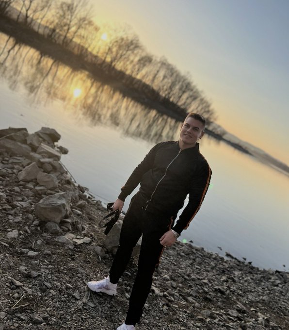 A man in a black tracksuit with rainbow-striped sleeves stands on a rocky shore beside a calm body of water at sunset, holding a camera strap.