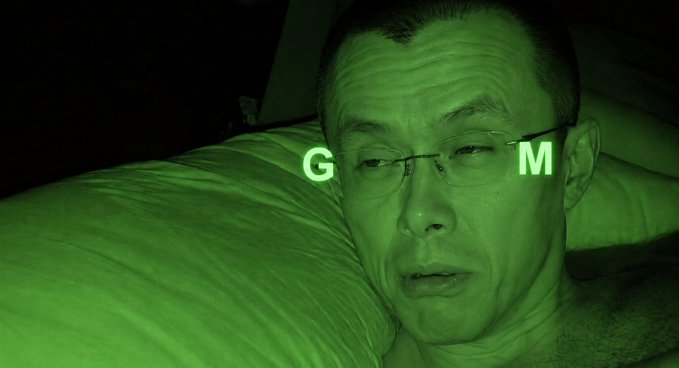 A close-up selfie of a man with glasses in low light with a green tint, with the letters 'G' and 'M' overlaid on the image.