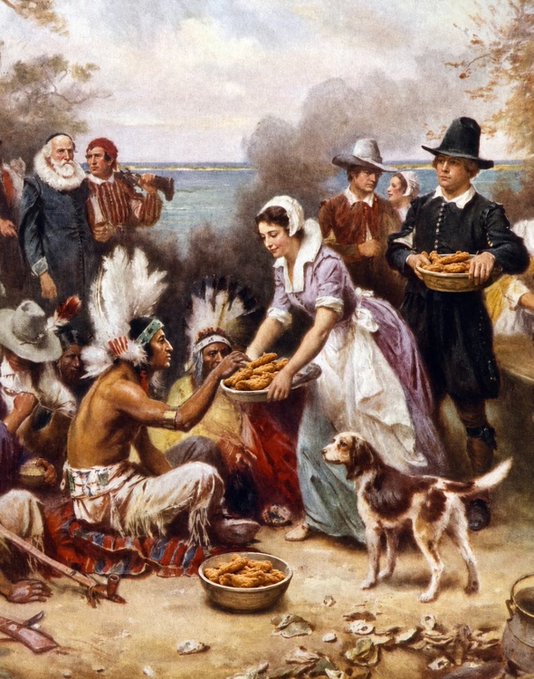 A historical painting depicting the First Thanksgiving, showing Pilgrims in traditional colonial attire sharing food with Native Americans wearing feathered headdresses on a shoreline.
