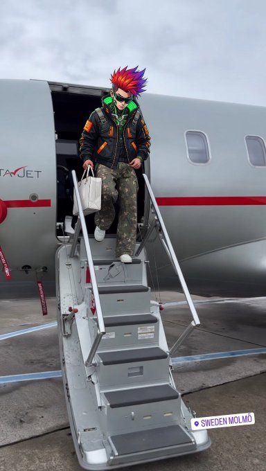 A person with bright red and purple hair wearing headphones, a black jacket, sunglasses, and camouflage pants descends the stairs of a private jet labeled "SWEDEN MOLMÖ" while carrying a small bag.