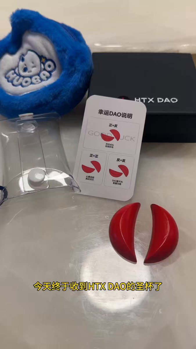 The image features a pair of red earrings with a white label that says "DAO" on them. The earrings are placed next to a blue stuffed animal hat, which has a cartoon character design. A black box containing a card is also present in the scene.