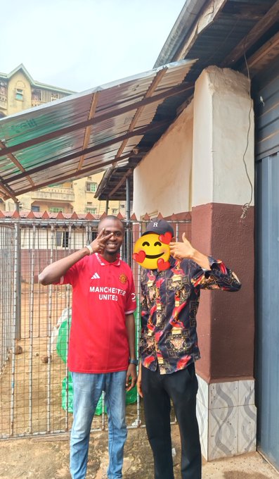 Two men pose together outside near a gate, with one wearing a red Manchester United jersey; the other person's face is covered by a kiss emoji sticker.