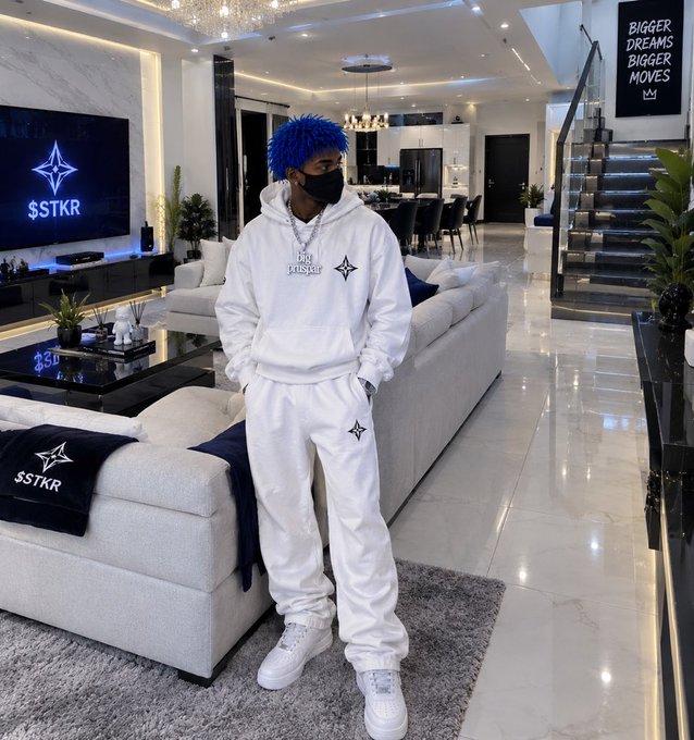A person with bright blue dreadlocks wearing a white hoodie and sweatpants set labeled 'big risker', posing in a modern luxury living room with 'STKR' branding and 'Bigger Dreams Bigger Moves' signage.