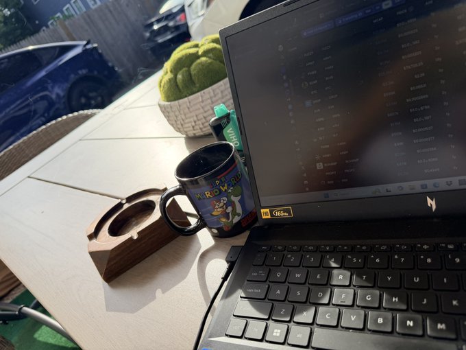 A laptop on a wooden table displays a spreadsheet with cryptocurrency values, next to a 'Happy Birthday' mug and a wooden coaster.