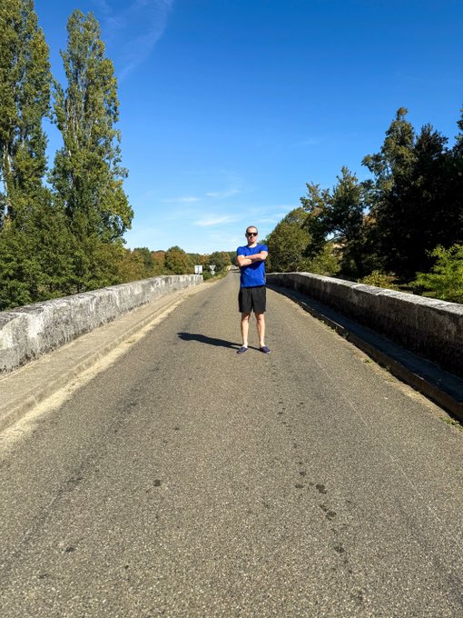 A man wearing a blue shirt and black shorts stands with crossed arms in the middle of a paved bridge on a sunny day, with trees and a blue sky in the background.