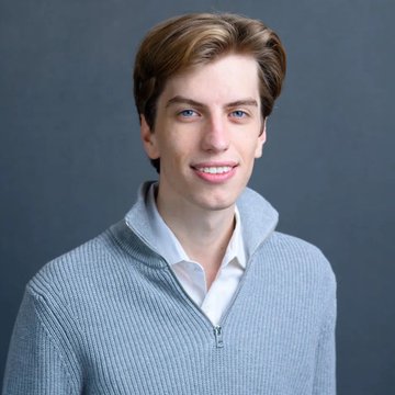 A young man with light brown hair smiling at the camera, wearing a white collared shirt under a gray ribbed zip-up sweater against a dark gray background.