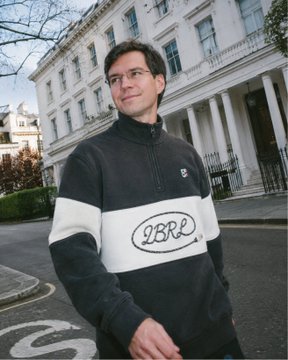 A young man stands on a London street wearing a black, white, and grey quarter-zip sweatshirt featuring the "2BR2" logo, with elegant white Georgian townhouses in the background. The image humorously illustrates the post's joke about the DoubleZero project "showing up" to BP (British Petroleum) wearing a London-style jumper, implying a casual approach to improving Solana's transaction speeds.