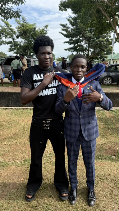Two young men pose outdoors on a grassy lawn, one in a black T-shirt reading “ALL … AMEBI” and the other in a blue checked suit with a red-and-blue sash, both making hand gestures. The image matches the post about a special appearance at a convocation, with Day 1 from @0xwale.