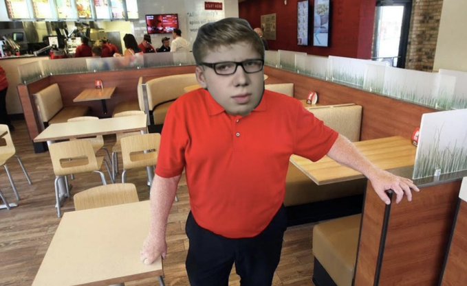 A young man wearing glasses and a red polo shirt standing in a fast-food restaurant booth area with light wood tables and chairs.