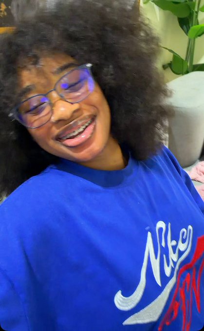 A smiling person with curly hair and glasses wearing a blue Nike shirt, with 'Nike' visible in white and red lettering.