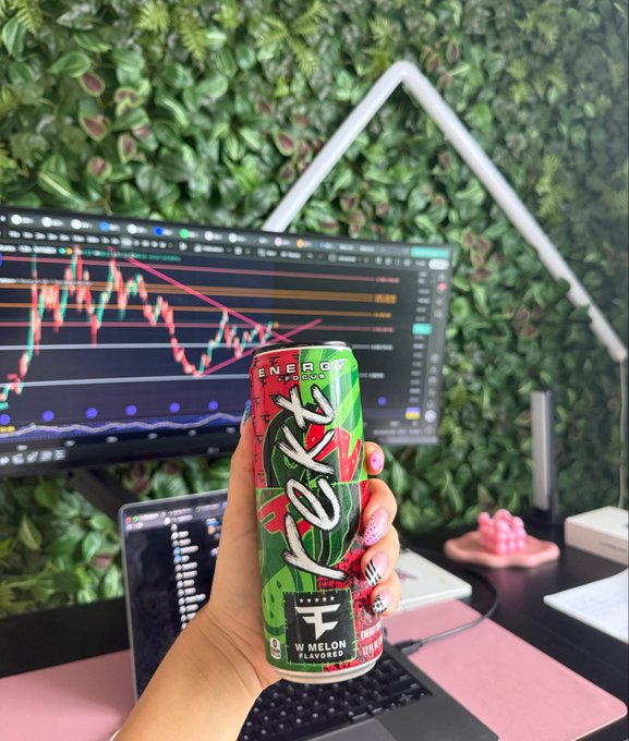 A hand with pink nail polish holds up a green and red 'VEX ENERGY' can (watermelon flavored) in front of a computer monitor displaying a trading chart, with a green plant wall in the background.
