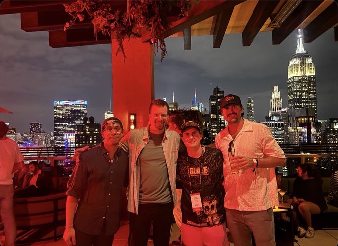 A group of four men posing together on a rooftop at night, with the illuminated New York City skyline, including the Empire State Building, in the background.
