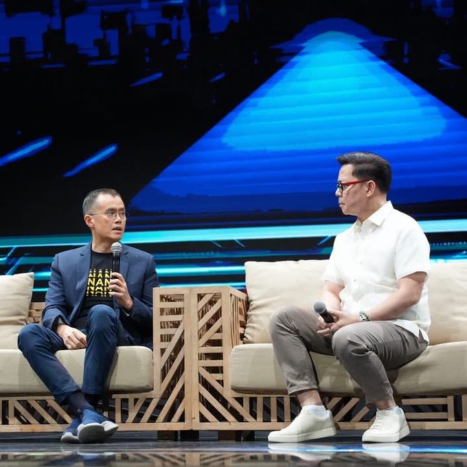 Two men sit on wooden crate-style chairs on a stage during what appears to be a panel discussion, with a large blue triangular screen in the background; one man wears a black t-shirt with 'NAN' visible.