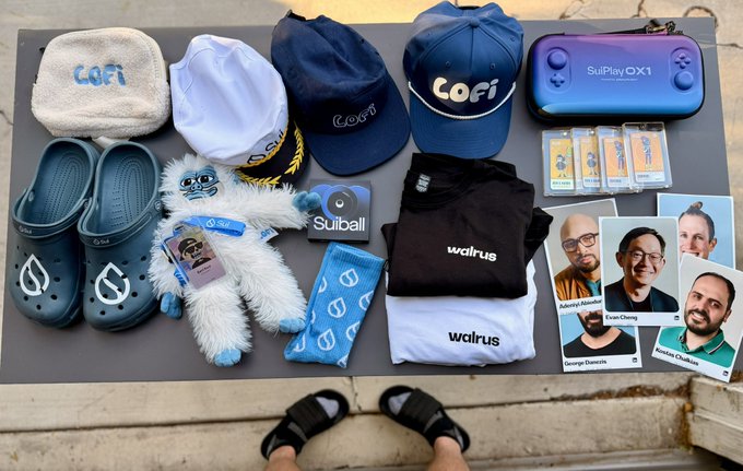 An overhead shot displaying branded merchandise including "LOFI" hats, "walrus" t-shirts, "SuiPlay OX1" device, "SuiBall" plushies, and headshots of four people labeled Aditya Chopra, Evan Cheng, George Danezis, and Kostas Chalkias.