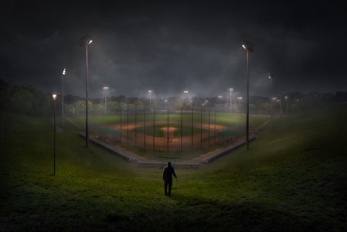 A lone figure stands in the foreground of a grassy field, gazing out at a baseball diamond illuminated by bright stadium lights cutting through the dark, moody night sky—evoking the solitary, atmospheric feeling of "Top of the Ninth," a baseball-themed NFT artwork by Hittman.