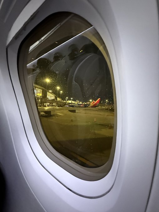 A nighttime view through an airplane window reveals the illuminated tarmac and another aircraft, likely taken while the plane is still at the gate. The image pairs with the post's tired "good night" message, capturing the quiet, weary moment of a traveler ready to finally rest after the flight.