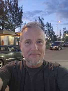 A selfie of a middle-aged man with gray stubble standing on a street at dusk, with cars, trees, and buildings visible in the background.