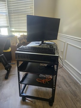 A black rolling AV cart holds a monitor, keyboard, and AV equipment in a room with wood flooring and white wainscoting.
