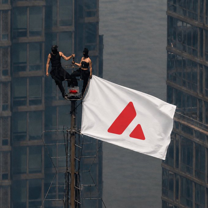 Two masked individuals sit atop an antenna displaying a white flag with a red Avax (Avalanche) logo, set against a city backdrop.
