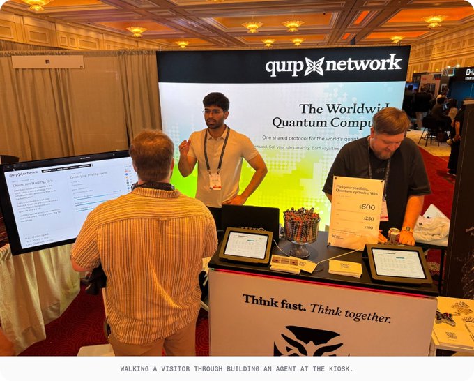 A photo of the Quip Network booth at a conference, where staff are demonstrating 'The Worldwide Quantum Computer' to a visitor, with the caption 'WALKING A VISITOR THROUGH BUILDING AN AGENT AT THE KIOSK.'