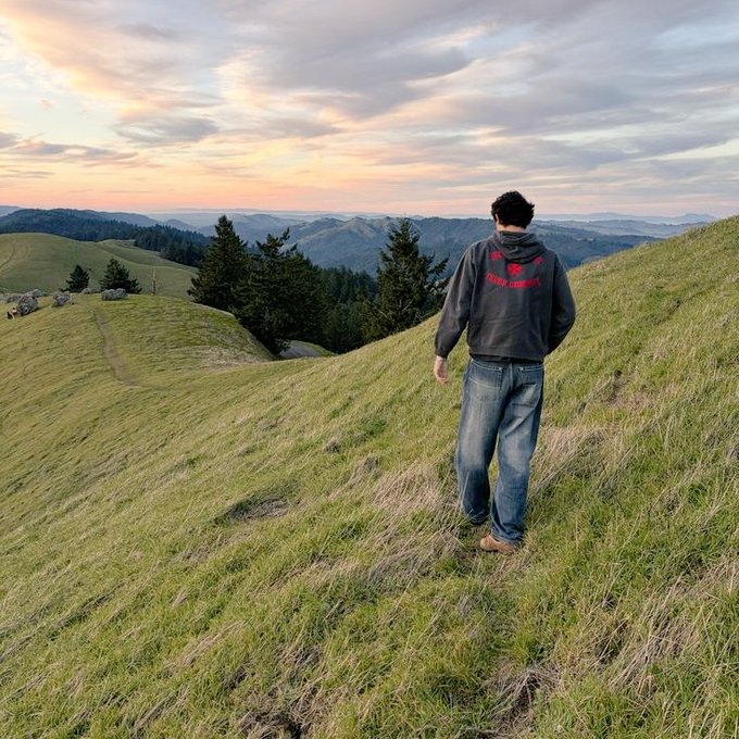 A man is walking through a grassy field, wearing jeans and a hoodie. The hillside he is traversing has several trees lining its slopes as well as cars parked nearby. There are also some small rocks visible in the area beneath his feet. He appears to be enjoying his walk up the side of the hill while taking in the beautiful view. In the image, there's a chain-abstraction design that looks like it could be related to River farming or another farming activity.