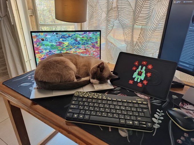 A cat curled up asleep on a closed laptop on a desk, next to a tablet displaying a green alien pixel game, with a mechanical keyboard and a second monitor in the background.