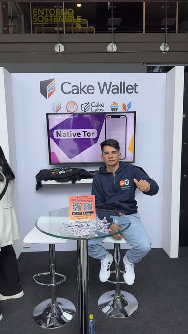 A young man in a black "Cake Labs" hoodie poses at the Cake Wallet booth at Criptolatinfest, standing next to a screen promoting "Native Tor" support and a table displaying promotional merchandise and QR codes for downloading the wallet app.