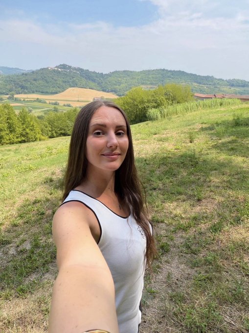A woman with long brown hair takes a selfie outdoors in a grassy field with rolling hills and farmland visible in the background.