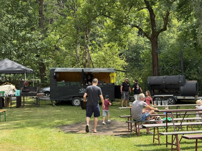 An outdoor gathering at a park features a black food truck, a smoker, and people sitting at picnic tables while a man walks with a child across the grass.
