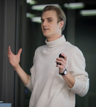 A young man in a cream turtleneck and wristwatch gestures while speaking, likely presenting or explaining a topic in a professional setting. His poised demeanor and engaging expression align with Fredrik Haga's entrepreneurial narrative, complementing the story of his academic and professional journey leading to the founding of Dune Analytics.