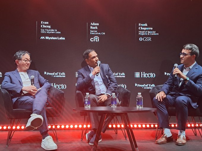 The image shows three men sitting around a table at an event, likely a panel discussion or conference. They are all wearing suits and appear to be engaged in conversation with each other. The background of the image features a stage setting with a microphone on it, suggesting that they might be discussing a topic related to technology or finance. 

There is also a bottle placed near one of the men's chair, possibly containing water or another beverage for them during the event.