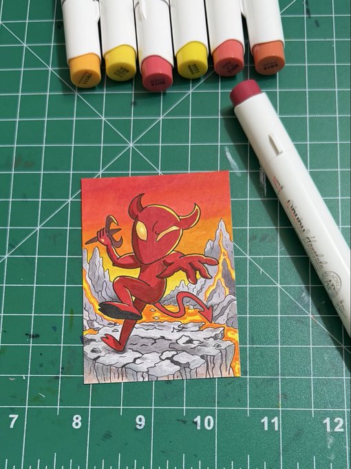 A small drawing of a red devil character with horns and a tail posing on rocks against a fiery background, sitting on a green cutting mat with alcohol markers above and beside it.