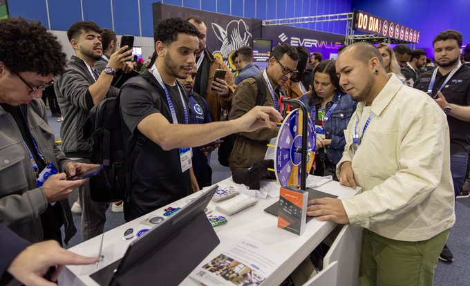 A photograph of people at a busy tech or gaming convention booth, with attendees examining products and one man in a yellow shirt speaking to the crowd.