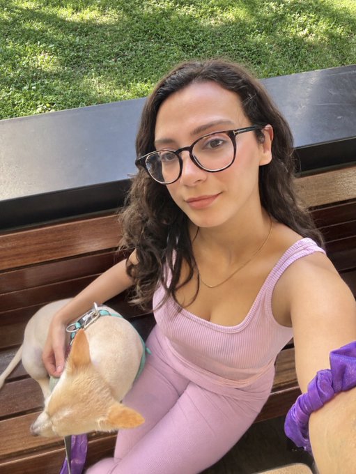 A woman with long dark hair and black-rimmed glasses takes a selfie while sitting on a wooden bench in a park, wearing a pink ribbed tank top and matching pants, with a small tan Chihuahua resting on her lap.